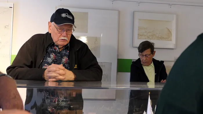 Ein älterer Mann mit Brille und schwarzer Kappe steht an einer Glasvitrine in einem Raum mit gerahmten Zeichnungen an der Wand.