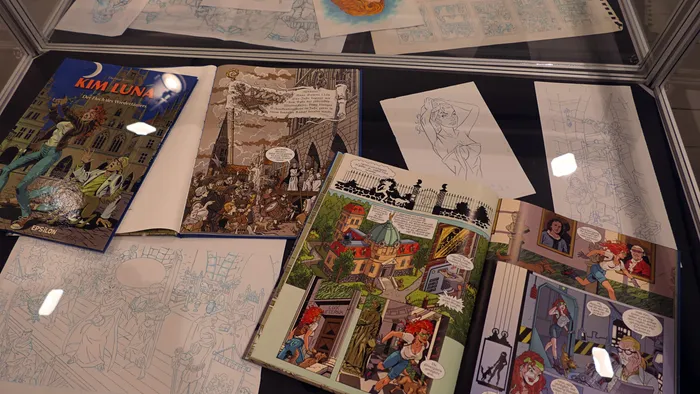 Verschiedene Comic-Bücher, Skizzen und aufgeschlagene Comic-Seiten in einer Glasvitrine.