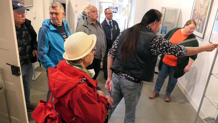 Ein Mann mit langen Haaren zeigt einer Gruppe von Besuchern in einem Ausstellungsraum ein Bild an der Wand.