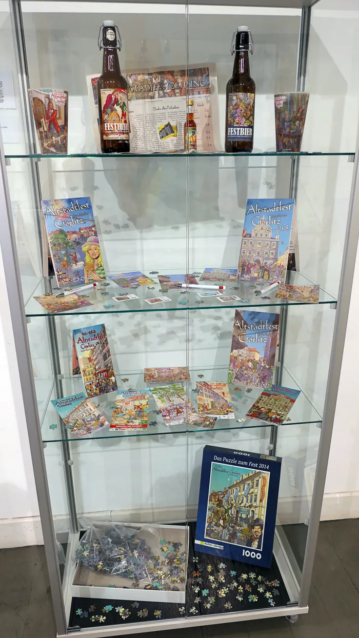 Glasvitrine mit verschiedenen Artikeln zum Altstadtfest Görlitz, darunter Bierflaschen, Broschüren, Postkarten und eine Puzzelbox.