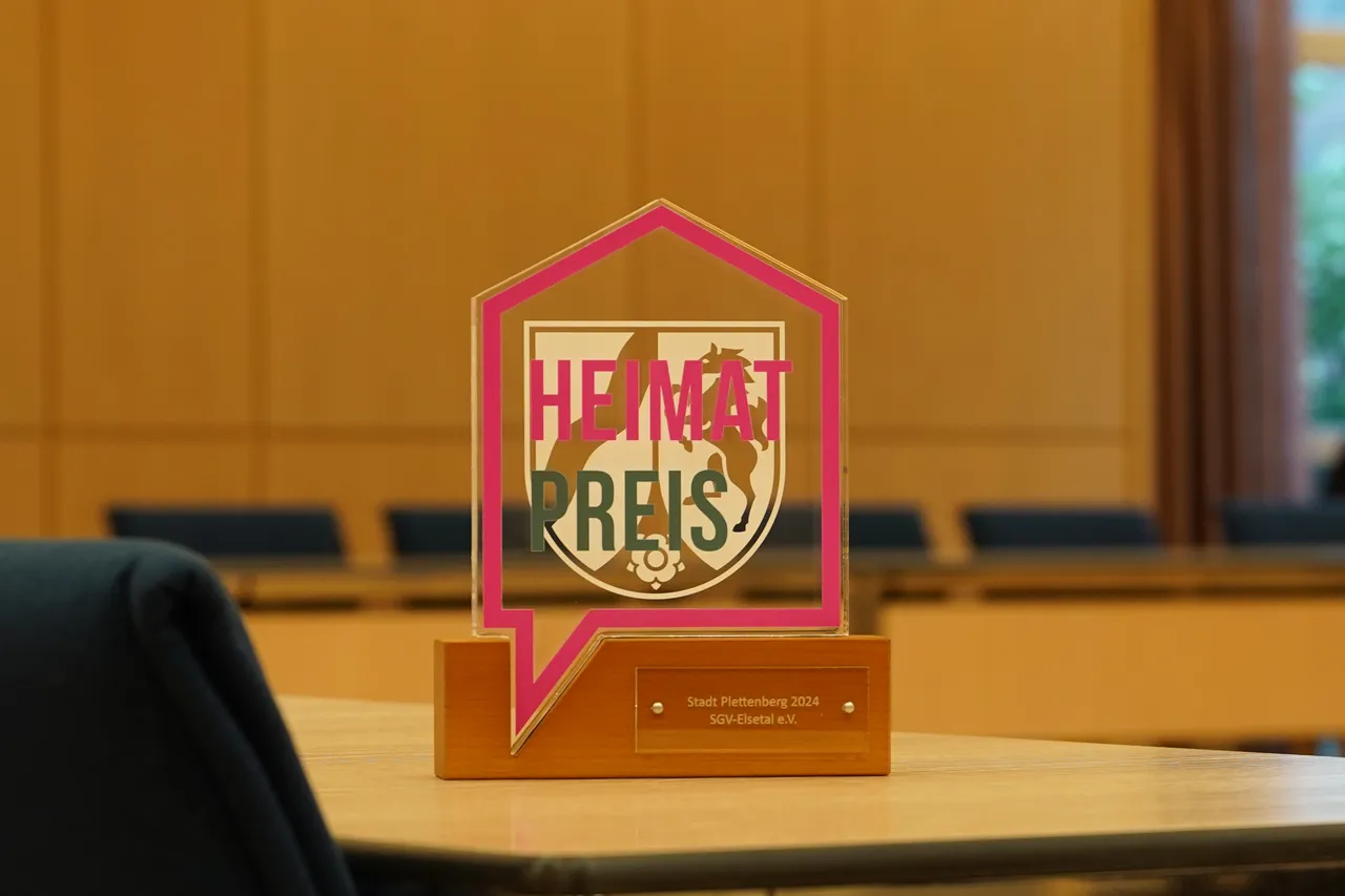 Der „Heimatpreis“ der Stadt Plettenberg 2024 ist auf einem gläsernen Sockel zu sehen. Auf dem Preis steht in großen roten Buchstaben „HEIMATPREIS“. Darunter befindet sich die Inschrift „Stadt Plettenberg 2024, SGV-Elsetal e.V.“