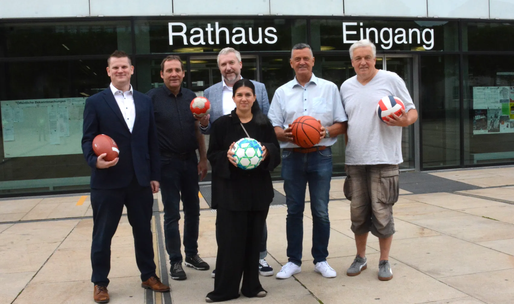 Sechs Personen, darunter Yasemin Kanca und Bürgermeister Wagemeyer, halten verschiedene Sportbälle vor dem Eingang des Rathauses.