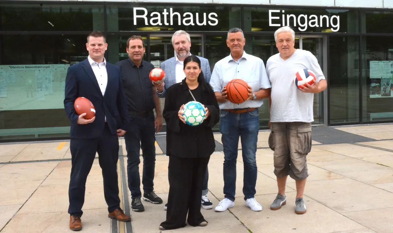 Sechs Personen stehen vor dem Eingang des Rathauses und halten verschiedene Sportbälle. Sie werben für den Heimatpreis 2026, der ehrenamtliches Engagement im Sport auszeichnet.