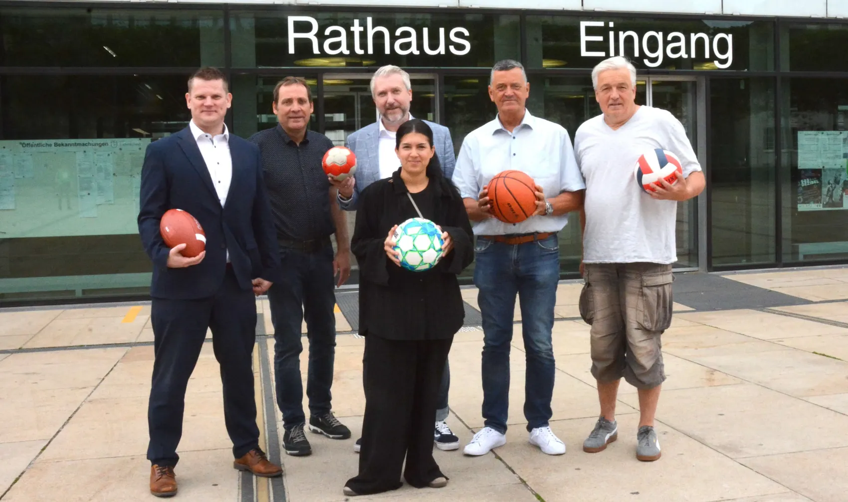 Sechs Personen stehen vor dem Eingang des Rathauses und halten verschiedene Sportbälle. Sie werben für den Heimatpreis 2026, der ehrenamtliches Engagement im Sport auszeichnet.