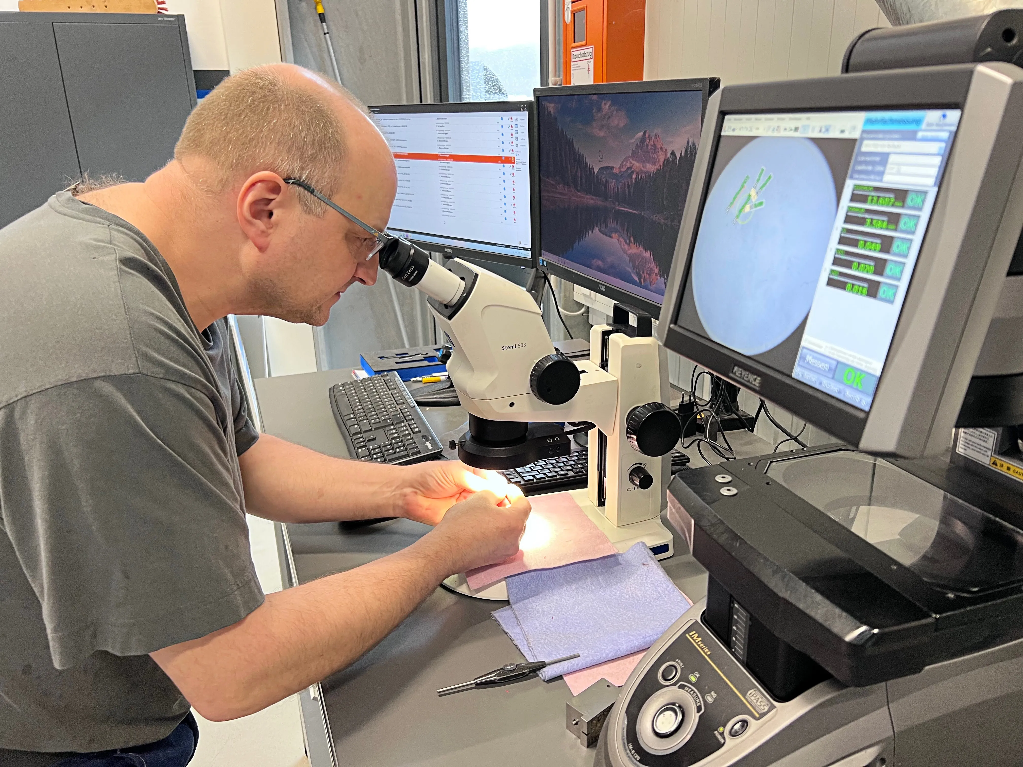 Ein Mann mit Kurzhaarfrisur arbeitet konzentriert mit einem Mikroskop und einem Computer an kleinen Metallteilen. Im Hintergrund sind weitere Monitore und technische Geräte zu sehen.