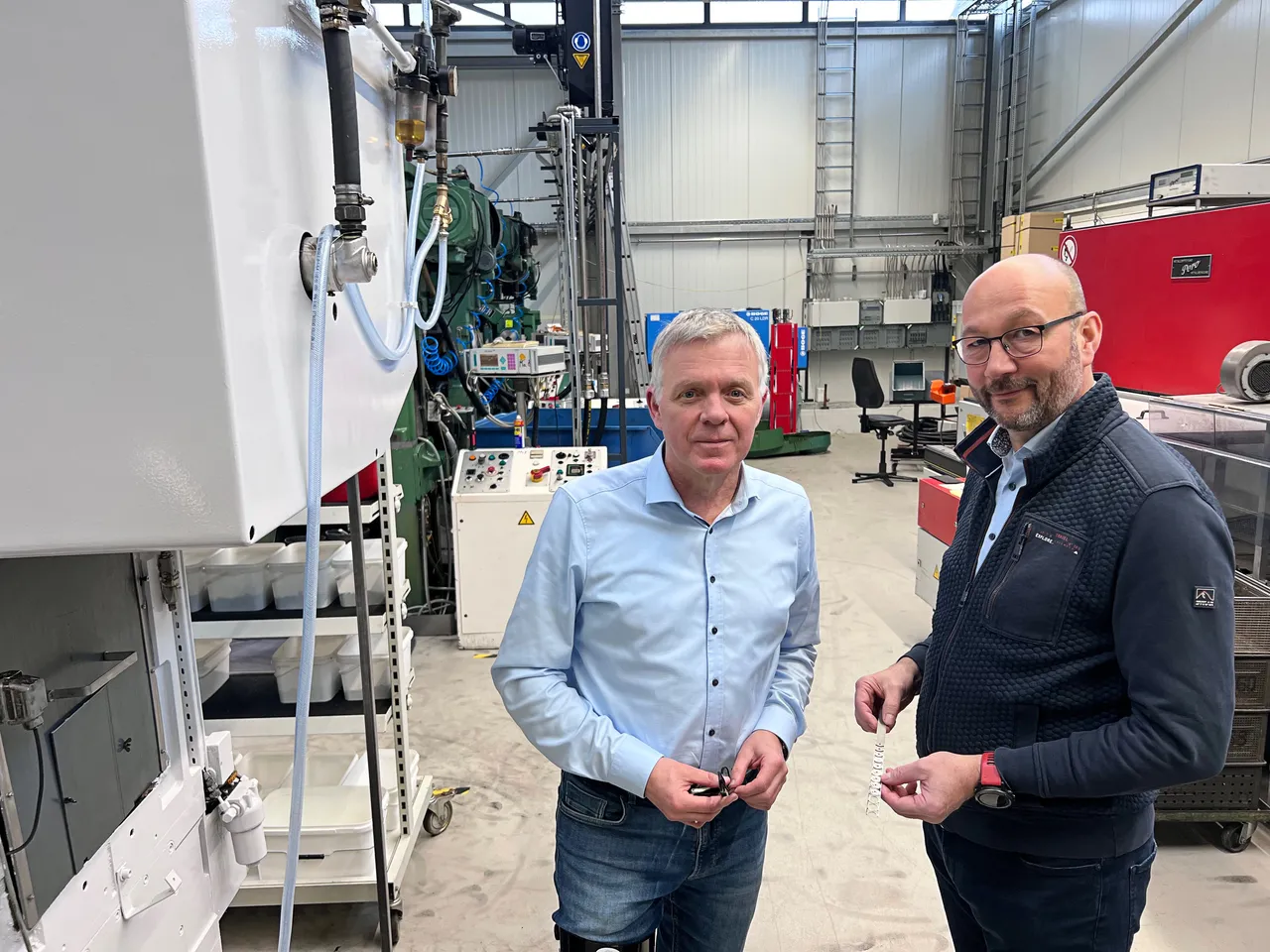 Zwei Männer stehen vor einer großen, hellen Maschine in einer Werkshalle. Der Mann links hält eine Metallfeder in der Hand, beide blicken in die Kamera.