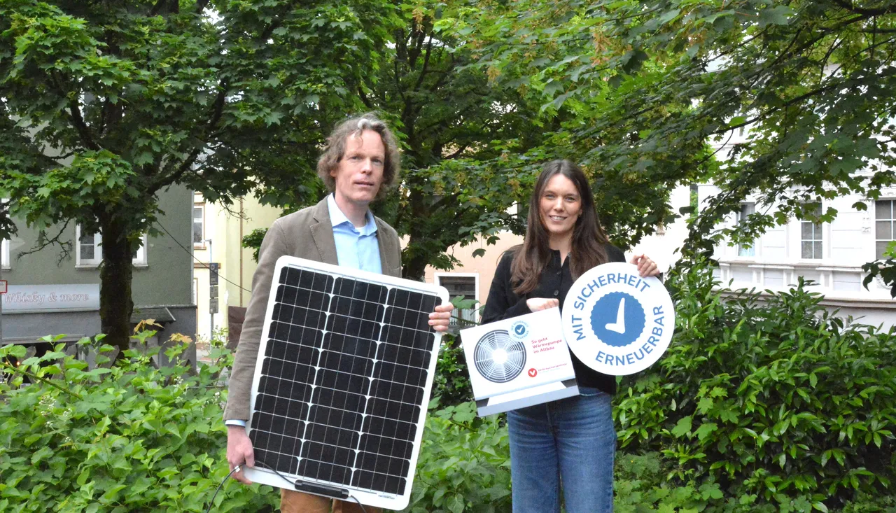 Ein Mann und eine Frau stehen lächelnd im Freien vor einem Gebäude. Der Mann hält ein Solarpanel, die Frau ein Schild mit der Aufschrift „Mit Sicherheit erneuerbar“.