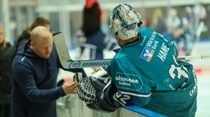 Eishockeytorwart Hendrik Hane in einem türkisfarbenen Trikot lehnt an der Bande, während ein Mann in einem dunkelblauen Kapuzenpullover zu ihm hinüberbeugt.