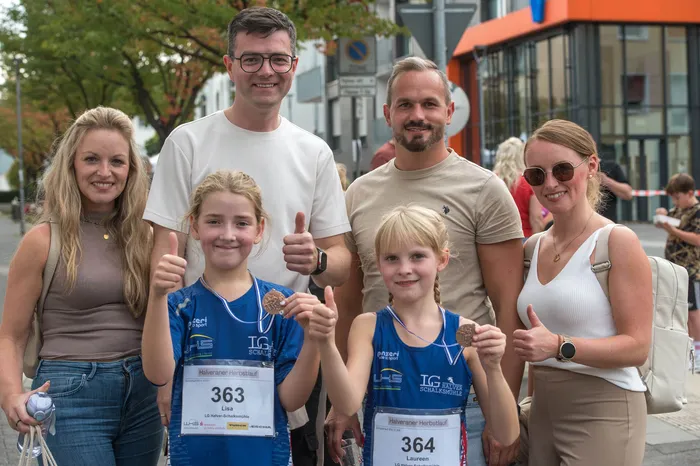 Zwei Mädchen mit Medaillen und Startnummern geben beim Halveraner Herbstlauf Daumen hoch, umgeben von einer Gruppe lächelnder Menschen.