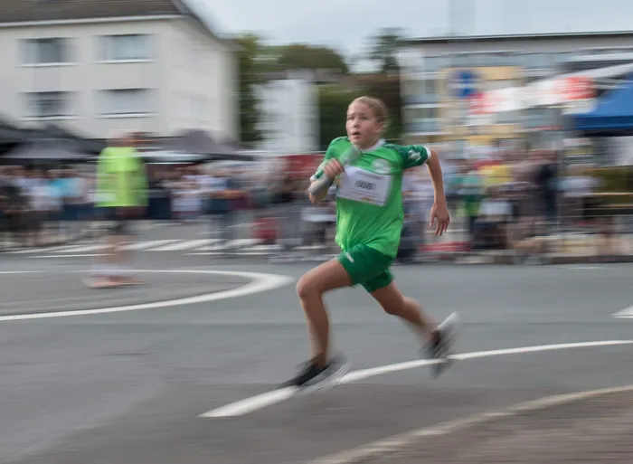 Eine junge Läuferin in grüner Sportkleidung rennt mit einem Staffelstab über eine Straße.