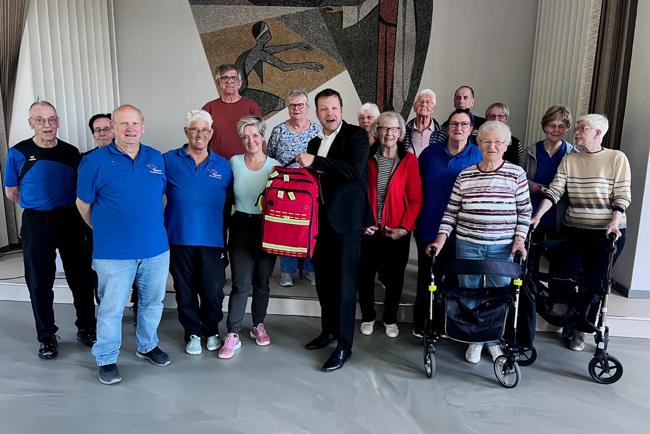 Gruppe von etwa zwanzig älteren Menschen steht vor einer Wand mit einem abstrakten Wandbild. In der Mitte hält ein Mann einen roten Notfallrucksack, überreicht von einem Vertreter der Vereinigten Sparkasse.