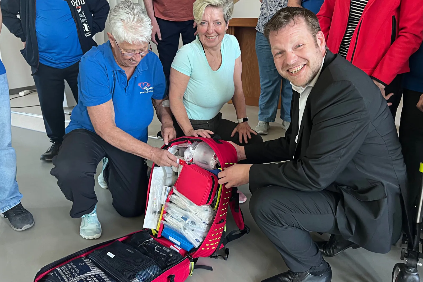 Zwei Frauen und ein Mann in formeller Kleidung betrachten den Inhalt eines geöffneten roten Notfallrucksacks mit medizinischer Ausrüstung.