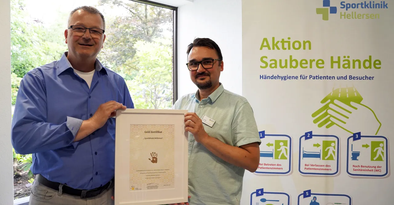 Zwei Männer halten ein Gold-Zertifikat der „Aktion Saubere Hände“ vor sich. Im Hintergrund ein Plakat mit Hinweisen zur Händehygiene.