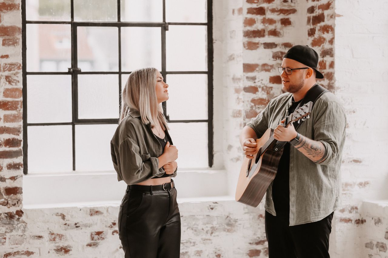 Ein Mann spielt Gitarre, während eine Frau ihm gegenübersteht und zuhört. Im Hintergrund ein großes Fenster mit Sprossen und eine Backsteinwand.
