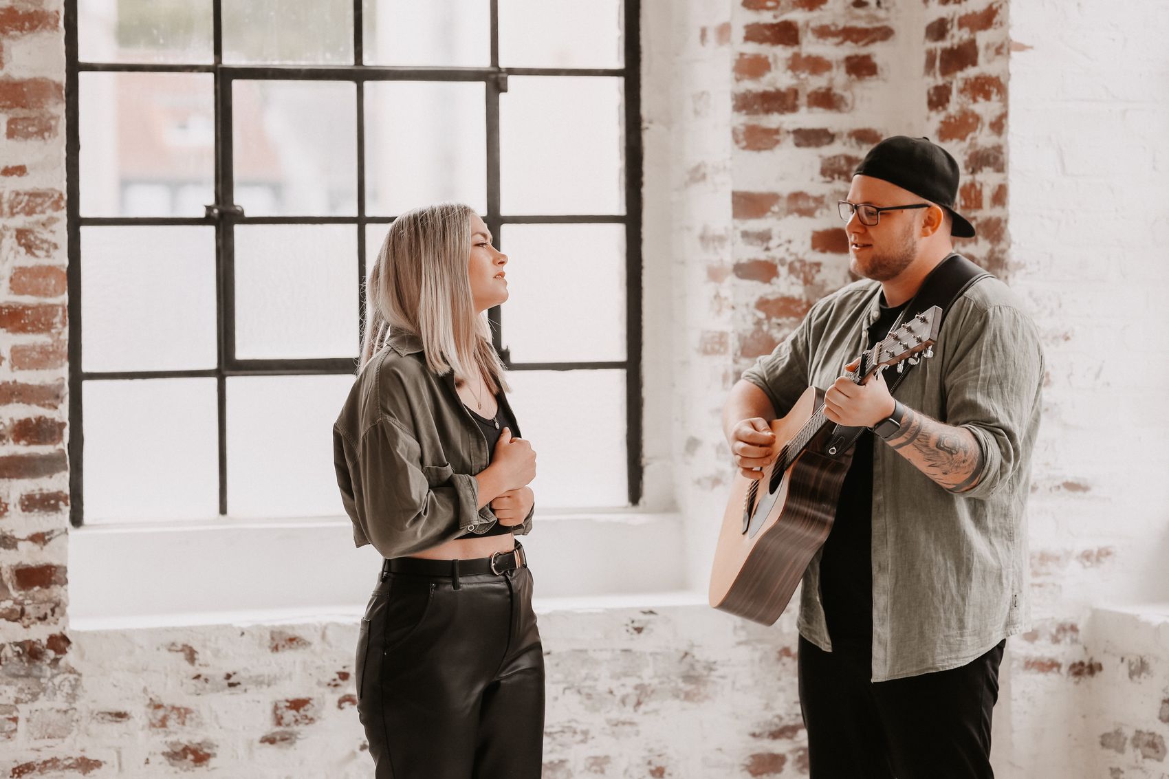 Ein Mann spielt Gitarre, während eine Frau ihm gegenübersteht und zuhört. Im Hintergrund ein großes Fenster mit Sprossen und eine Backsteinwand.