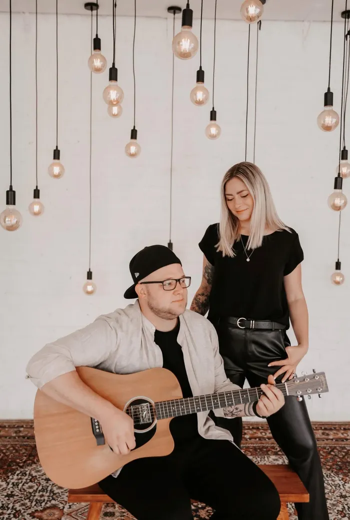 Ein Mann spielt Gitarre und eine Frau steht neben ihm vor einer Wand mit vielen hängenden Glühbirnen.