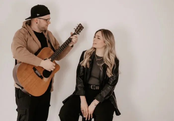 Ein Mann spielt Gitarre für eine Frau, die lächelnd auf einem Hocker sitzt. Im Hintergrund eine weiße Wand.
