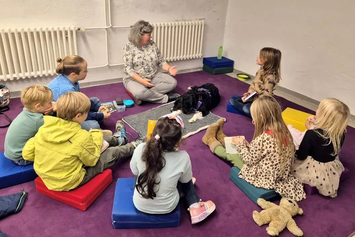 Anita Houben und ihr Hund Lara sitzen mit Kindern im Kreis auf bunten Kissen auf einem lila Teppich.