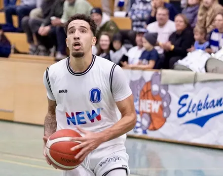Ein Basketballspieler mit dunkler Hautfarbe dribbelt den Ball während eines Spiels. Er trägt ein weißes Trikot mit den Aufschriften „NEW“ und „Jako“ sowie die Logos der „Iserlohn Roosters“ und „Rheingas“. Im Hintergrund sind verschwommene Zuschauer und das Logo „Elephants“ zu sehen.