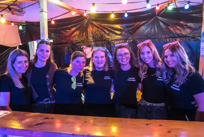 Gruppe von jungen Frauen mit Accessoires wie Brillen und Diademen vor einer Bar, alle tragen T-Shirts mit Aufschrift „Feuerwehr Schalksmühle“.