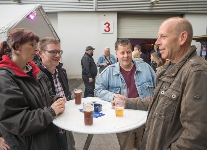 Vier Personen stehen um einen runden Bistrotisch mit Getränken und unterhalten sich angeregt. Im Hintergrund weitere Menschen und ein Gebäude mit der Nummer 3.