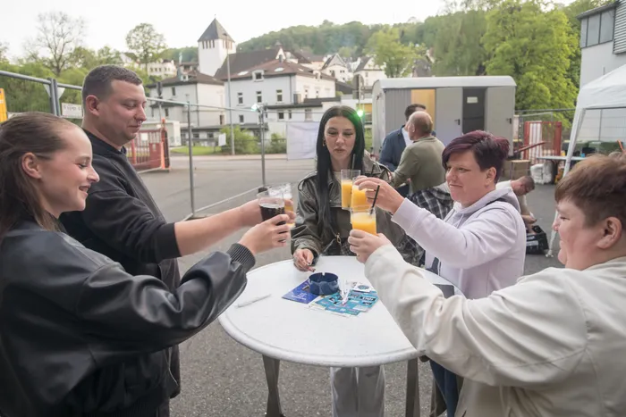 Fünf Personen stoßen mit Getränken an einem runden Tisch im Freien an, im Hintergrund ein Gebäude und weitere Menschen.
