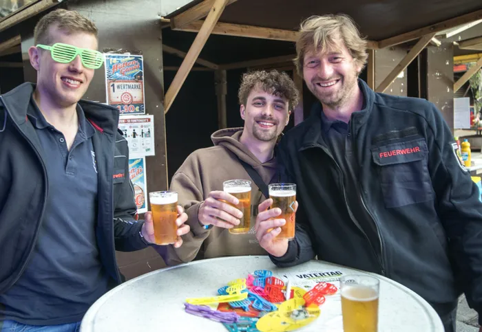 Drei junge Männer lächeln in die Kamera und heben jeweils ein Bierglas. Im Hintergrund ein Plakat mit der Aufschrift „Vatertag“ und ein Schild mit der Aufschrift „Feuerwehr“.