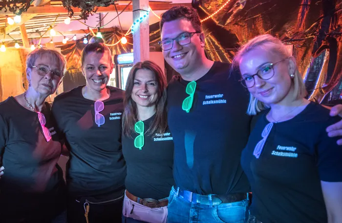 Fünf lächelnde Menschen vor einer Bar tragen schwarze T-Shirts mit dem Aufdruck „Feuerwehr Schalksmühle“ und einem stilisierten Feuerwehrauto.