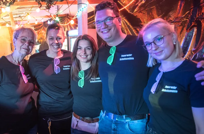 Fünf lächelnde Menschen stehen eng zusammen vor einer rustikalen Holzwand mit Lichterketten. Sie tragen schwarze T-Shirts mit dem Aufdruck „Feuerwehr Schalksmühle“ und einem stilisierten Feuerwehrmann.