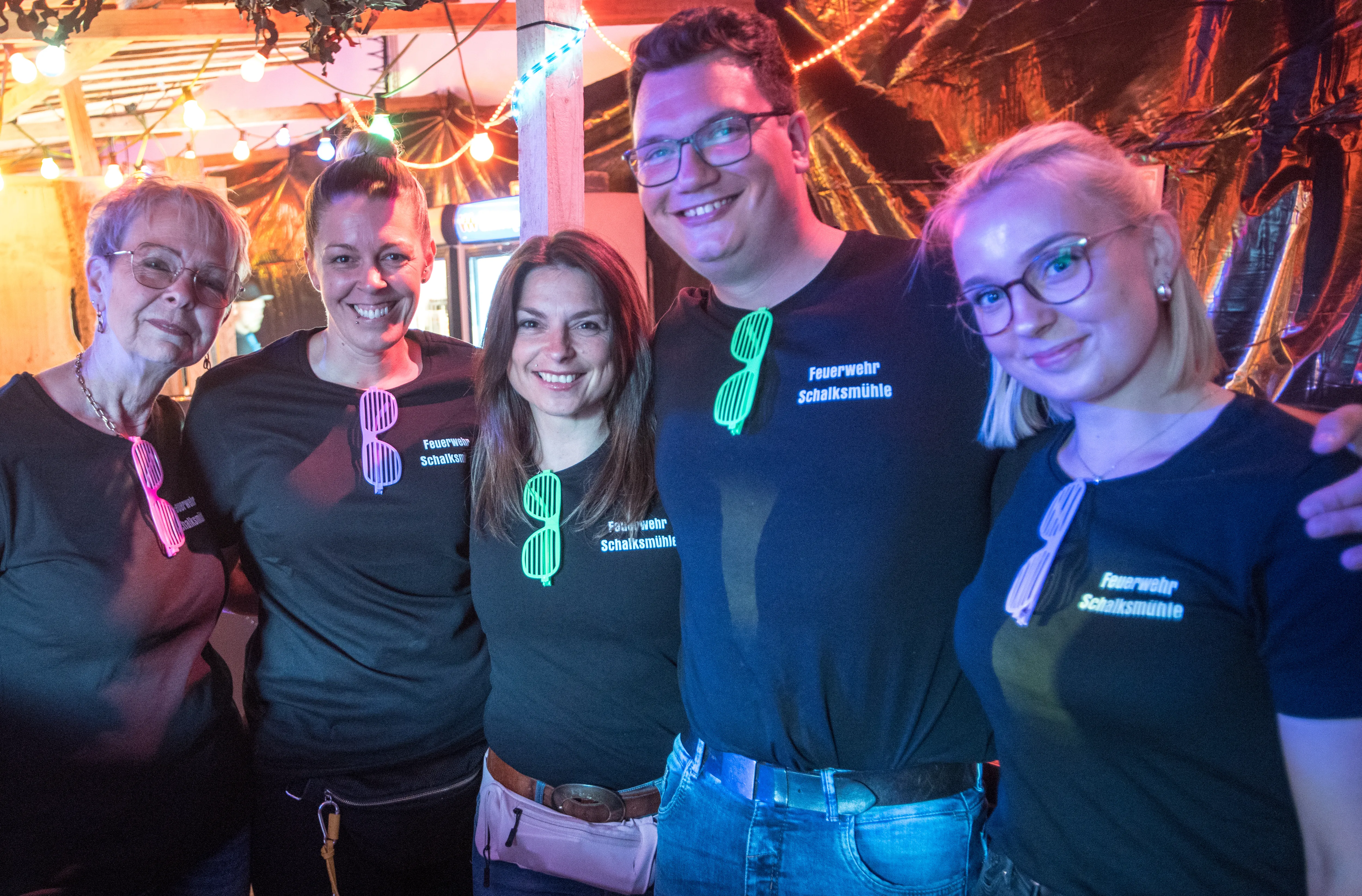 Fünf lächelnde Menschen stehen eng zusammen vor einer rustikalen Holzwand mit Lichterketten. Sie tragen schwarze T-Shirts mit dem Aufdruck „Feuerwehr Schalksmühle“ und einem stilisierten Feuerwehrmann.
