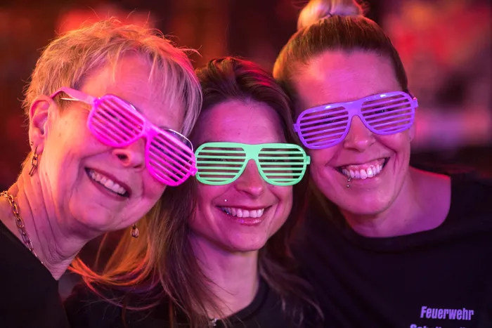 Drei lächelnde Frauen mit auffälligen Sonnenbrillen mit Lamellen vor rotem Hintergrund.