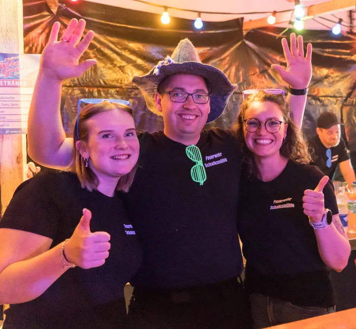 Drei lächelnde Menschen vor einer Bar mit Getränkekarte, zwei Frauen und ein Mann mit Cowboyhut, alle tragen T-Shirts mit dem Aufdruck „Feuerwehr Schalksmühle“ und zeigen Daumen hoch oder winken.