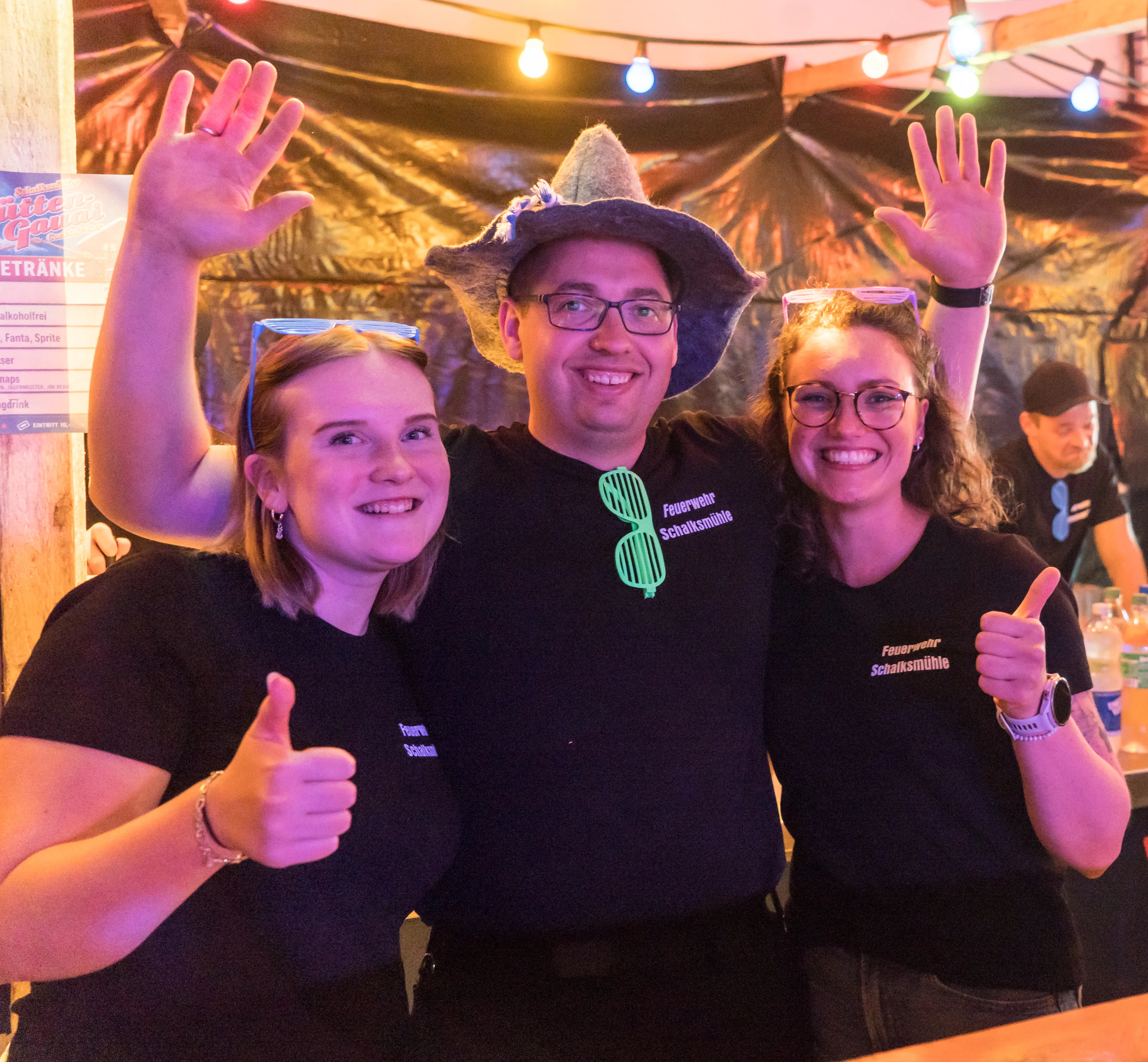 Drei lächelnde Menschen vor einer Bar mit Getränkekarte, zwei Frauen und ein Mann mit Cowboyhut, alle tragen T-Shirts mit dem Aufdruck „Feuerwehr Schalksmühle“ und zeigen Daumen hoch oder winken.