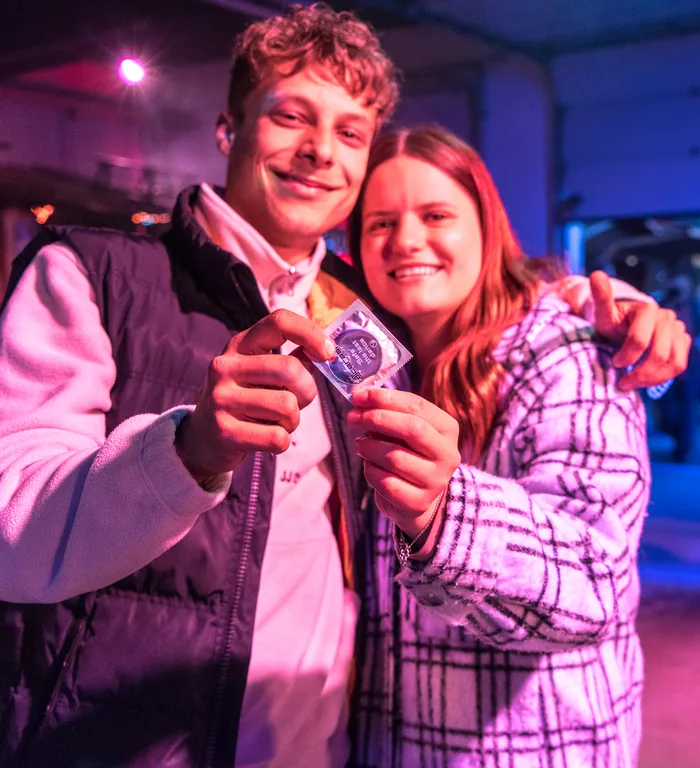 Ein junger Mann und eine junge Frau halten gemeinsam ein Kondom in die Kamera. Sie lächeln und stehen eng umschlungen vor einer verschwommenen Hintergrundkulisse.
