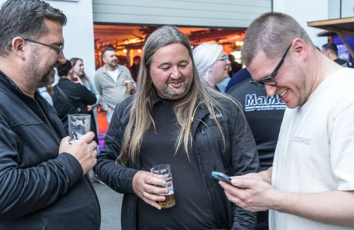 Drei Männer unterhalten sich auf der Hüttengaudi im Feuerwehrgerätehaus Schalksmühle. Ein Mann mit langen Haaren hält ein Glas Bier und schaut lächelnd zu den anderen beiden, die ein Smartphone betrachten.