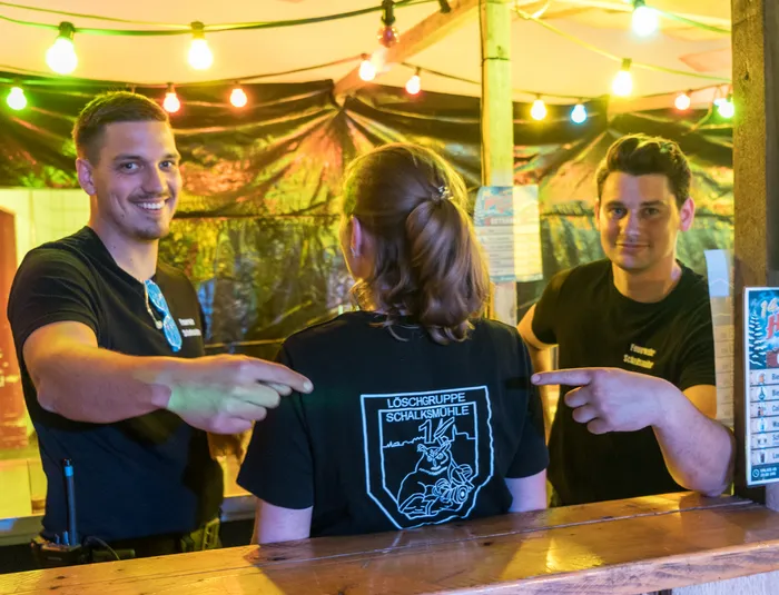 Zwei Männer lächeln und zeigen auf eine Person mit langem Haar, deren Rücken dem Betrachter zugewandt ist. Auf dem T-Shirt der Person steht „Löschgruppe Schalksmühle“.