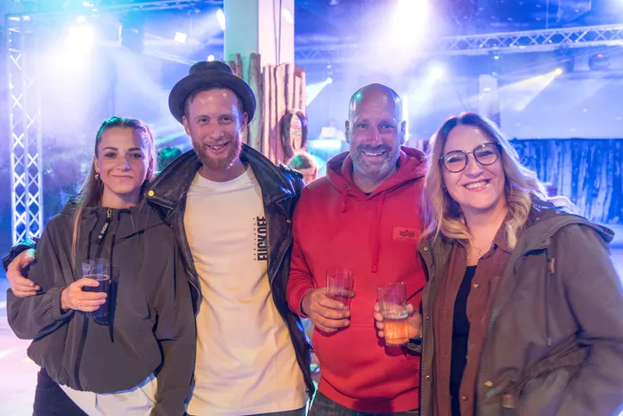 Vier lächelnde Menschen stehen eng zusammen und halten Gläser in der Hand, im Hintergrund eine festlich beleuchtete Halle.