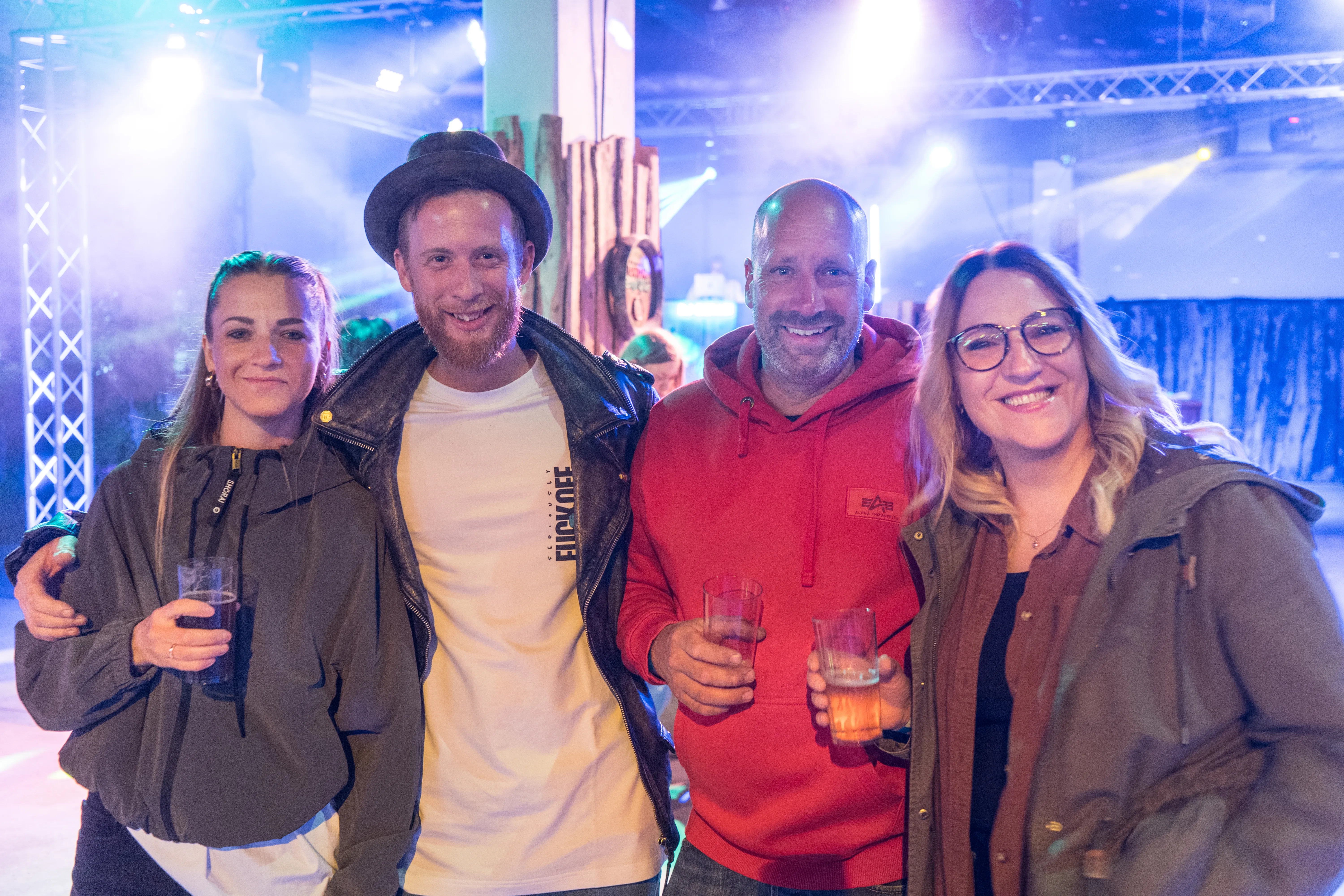 Vier lächelnde Menschen stehen eng zusammen und halten Gläser in der Hand, im Hintergrund eine festlich beleuchtete Halle.