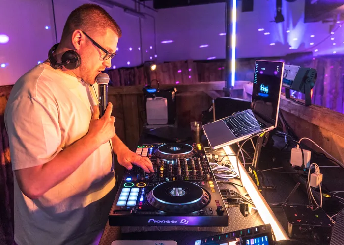 Ein DJ mit Kopfhörern bedient ein Mischpult der Marke Pioneer DJ vor Publikum in einem abgedunkelten Raum. Rechts steht ein Laptop auf einem Tisch.