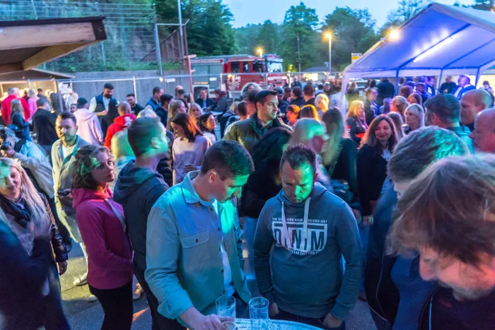 Eine große Menschenmenge feiert im und vor einem Feuerwehrgerätehaus. Im Vordergrund stehen zwei Männer, die sich unterhalten, während im Hintergrund viele weitere Besucher zu sehen sind.