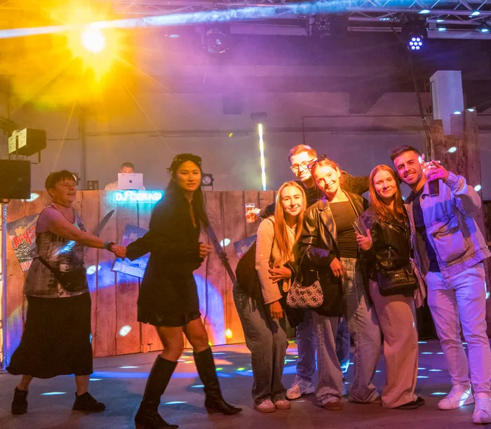 Eine Gruppe junger Erwachsener steht vor einer DJ-Anlage in einem Raum mit gedämpftem Licht und bunten Scheinwerfern. Im Hintergrund ist ein DJ zu sehen, der an einem Laptop arbeitet.