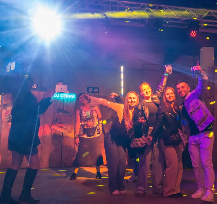 Eine Gruppe junger Erwachsener feiert in einem beleuchteten Raum, vermutlich in einem Feuerwehrgerätehaus, mit tanzenden Personen und einem DJ-Pult im Hintergrund.