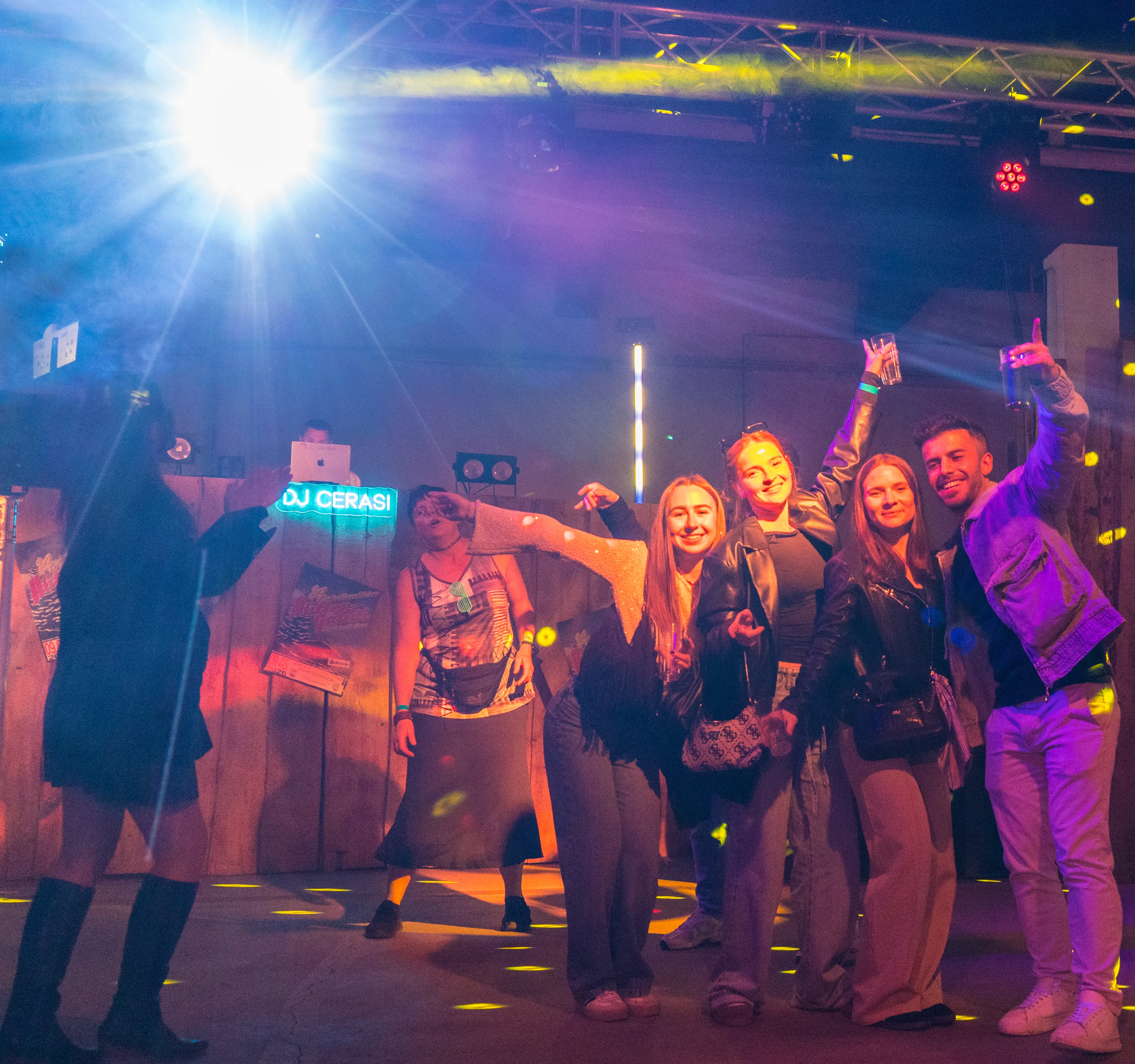 Eine Gruppe junger Erwachsener feiert in einem beleuchteten Raum, vermutlich in einem Feuerwehrgerätehaus, mit tanzenden Personen und einem DJ-Pult im Hintergrund.