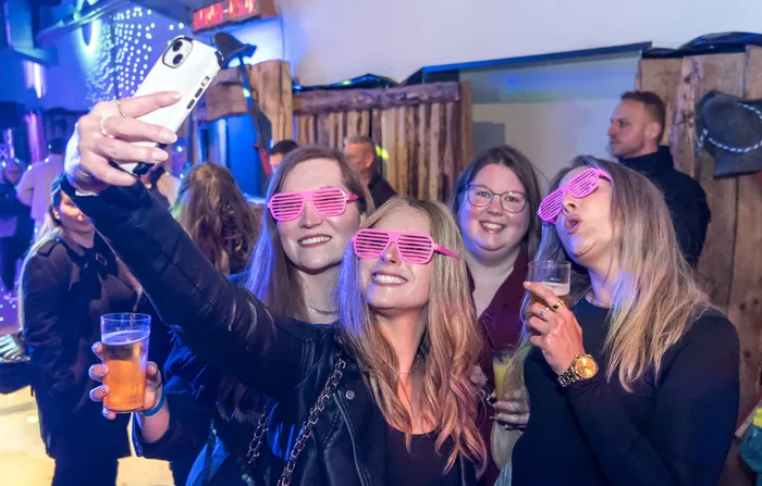 Eine Gruppe junger Frauen macht ein Selfie auf einer Party, alle tragen lustige Sonnenbrillen und halten Getränke in der Hand.