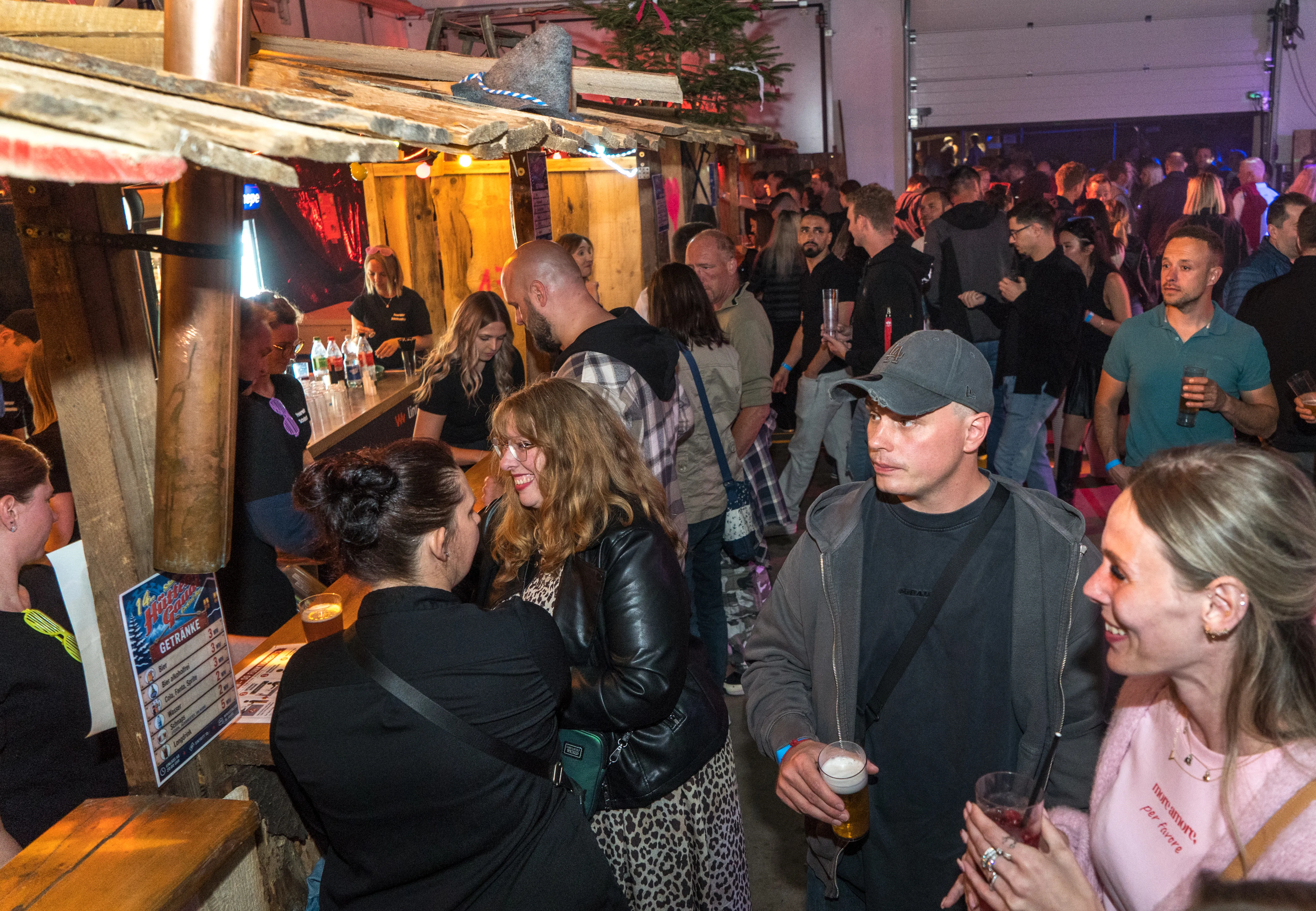Menschen stehen und unterhalten sich an einer Bar in einem voll besetzten Raum, vermutlich auf einem Fest im Feuerwehrgerätehaus.