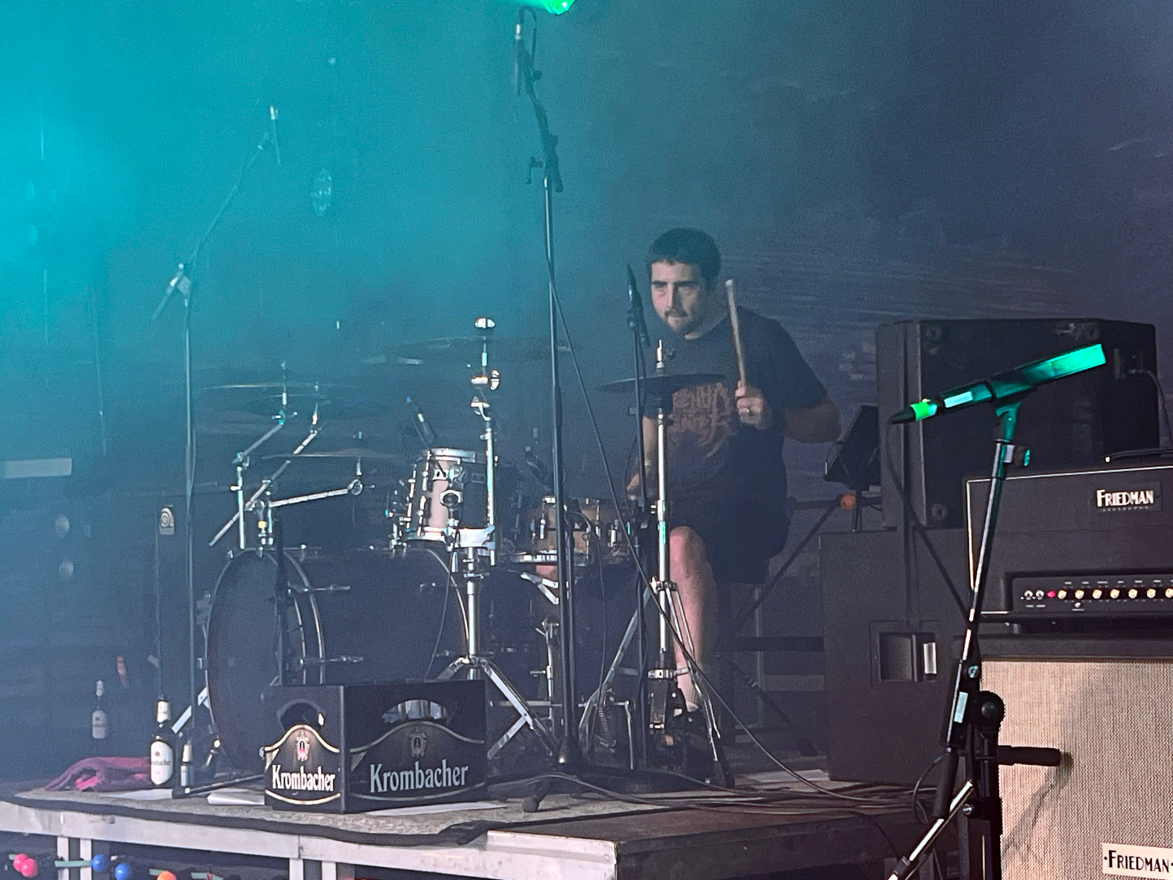 Schlagzeuger spielt auf einer Festivalbühne. Im Hintergrund sind Verstärker und Bühnenbeleuchtung zu sehen, vor der Bühne stehen zwei Bierfässer mit dem Aufdruck „Krombacher“.
