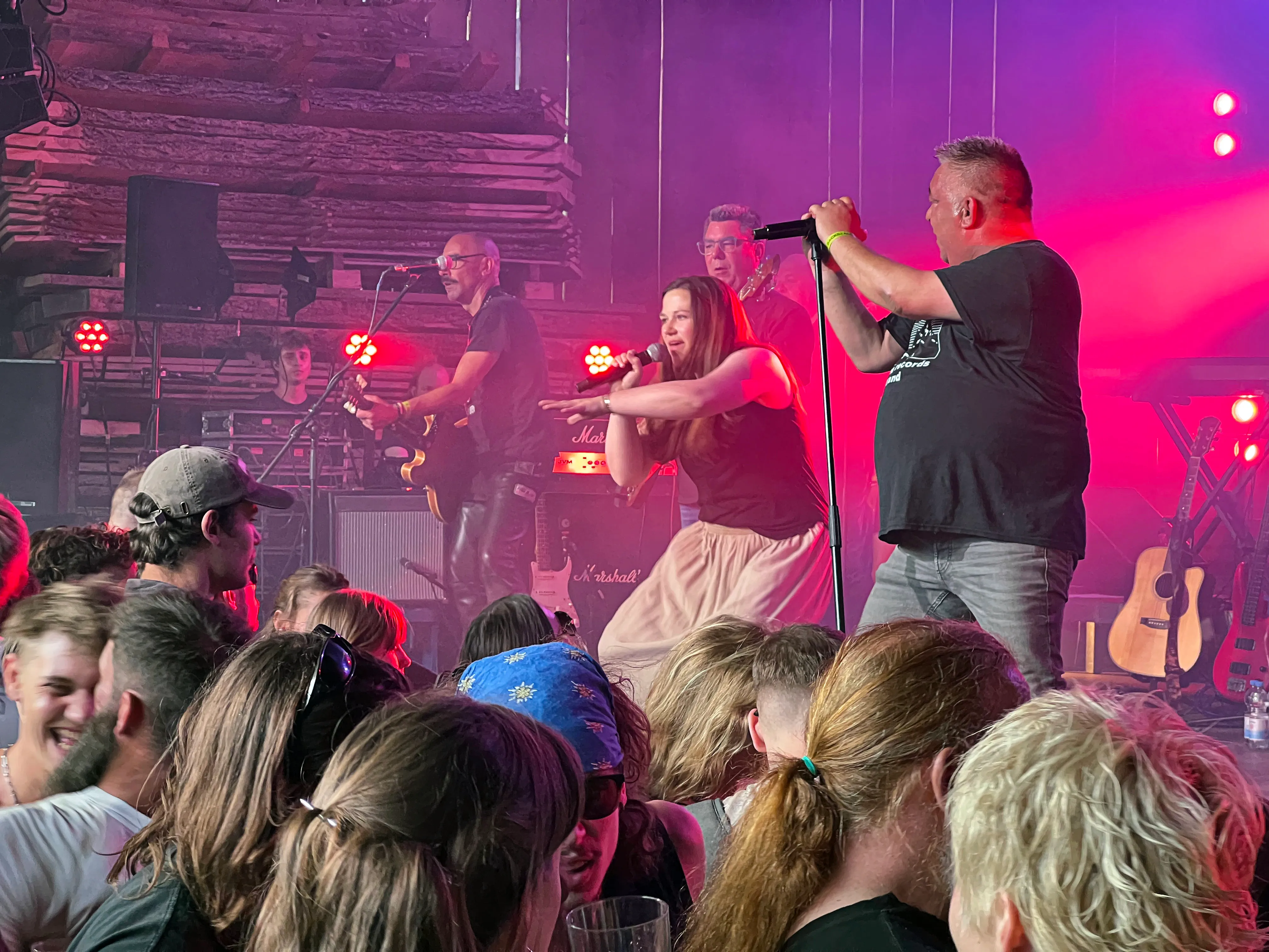 Eine Band spielt vor einem Publikum im Freien. Ein Sänger mit Mikrofon steht rechts im Vordergrund, während eine Sängerin links davon singt und ein Gitarrist im Hintergrund zu sehen ist.