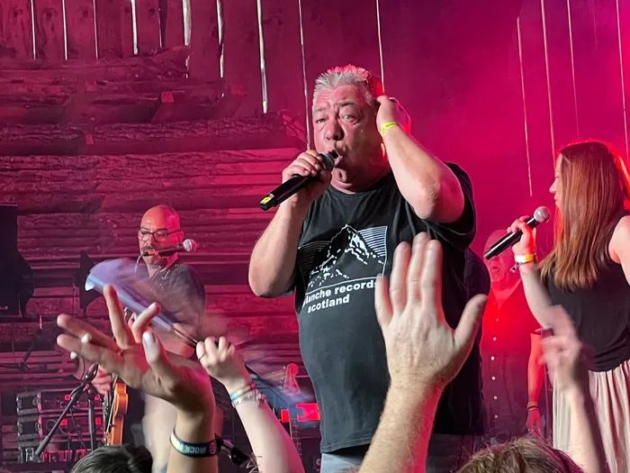 Zwei Sänger performen auf der Bühne des I-Rock Festivals, während das Publikum die Hände hebt. Im Hintergrund sind weitere Musiker und eine rustikale Holzwand zu sehen.