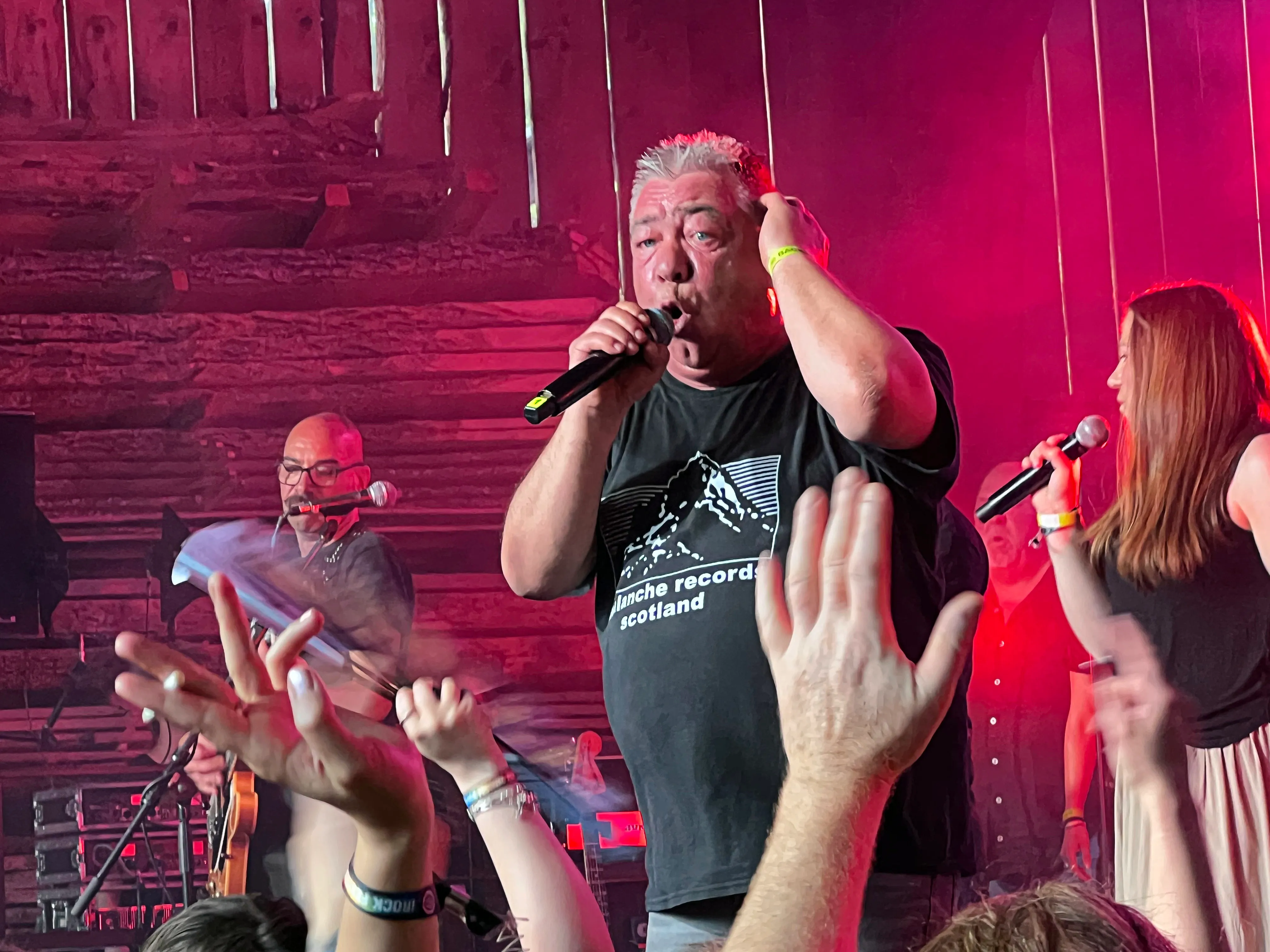 Zwei Sänger performen auf der Bühne des I-Rock Festivals, während das Publikum die Hände hebt. Im Hintergrund sind weitere Musiker und eine rustikale Holzwand zu sehen.
