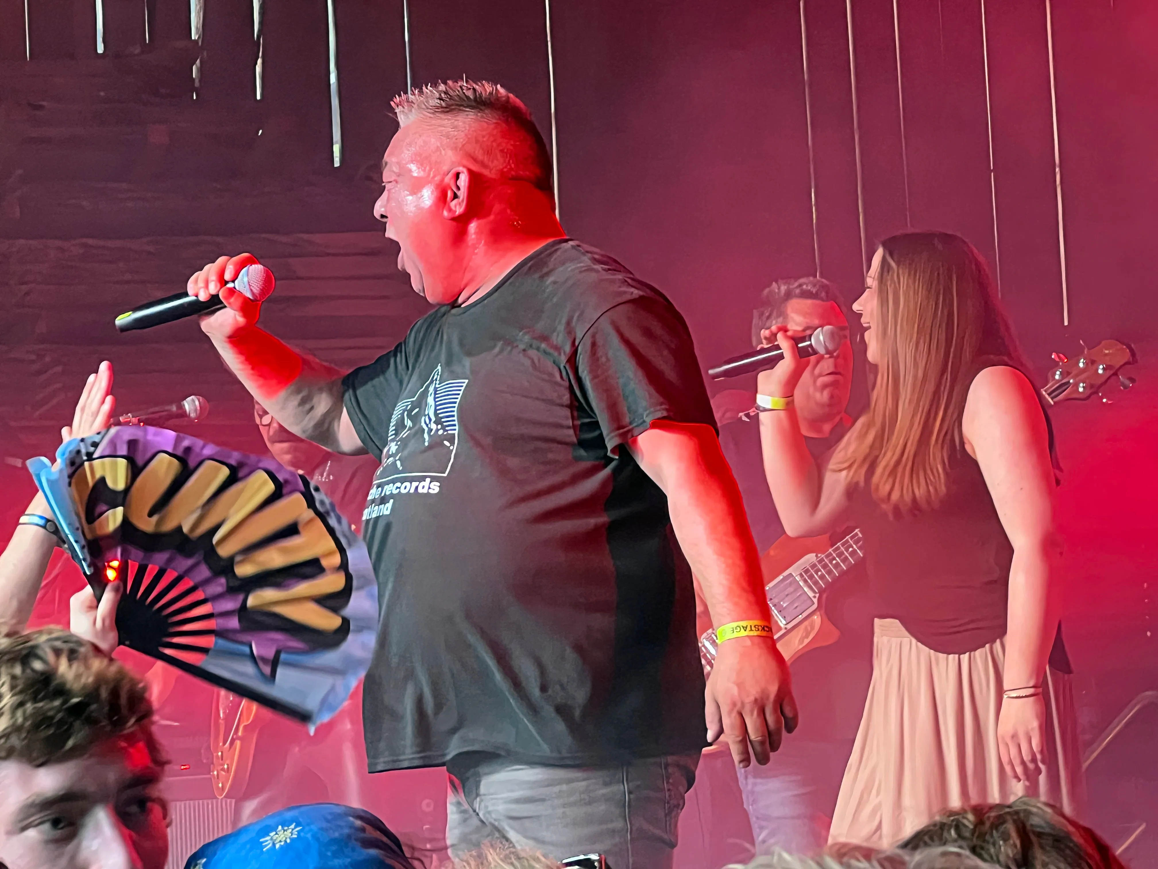 Zwei Musiker auf der Bühne des I-Rock Festivals, ein Mann singt in ein Mikrofon, eine Frau hält ein weiteres Mikrofon. Im Vordergrund eine Person mit einem Fächer.